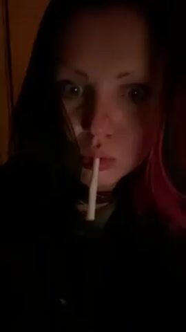 A woman with dark hair and pink highlights is lighting a cigarette held in her mouth. She is holding a lighter in her left hand which is covered in tattoos and partially wrapped in clear plastic. She looks directly at the camera, her eyes are visible. The background is dark, but a doorframe can be seen to her left.