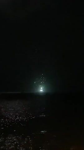 The video captures a nighttime scene at a body of water, likely the ocean, with a dark sky and shimmering reflections on the water's surface. In the distance, a bright light, possibly from a vehicle or a stationary source, is visible on what appears to be a pier or a land structure extending into the water. Periodically, bursts of fireworks illuminate the sky above the distant light. One firework, with a red hue, appears briefly in the upper portion of the frame. The overall ambiance is dark and atmospheric, with the intermittent explosions of fireworks providing flashes of light against the night sky.