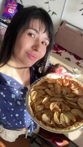 A woman with black hair and bangs smiles at the camera, holding a freshly baked apple pie. She is wearing a blue patterned shirt and a black choker necklace. The pie is in a glass dish and is arranged with thin slices of apple in a circular pattern. In the background, there is a table with several small bowls containing food, including a salad with red and green ingredients and a bowl with what appears to be nuts or crackers. The lighting suggests the scene is taking place indoors, possibly near a window, as evidenced by the strong sunlight casting shadows. The woman is looking directly at the camera, and the pie is positioned slightly to her right.