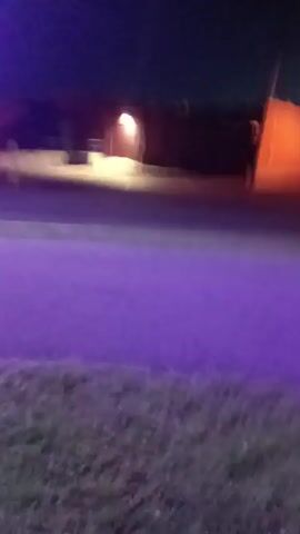 The video shows a dark scene with some purple light shining on a patch of grass in the foreground. In the background, there are some buildings and lights. The camera appears to be moving slightly.