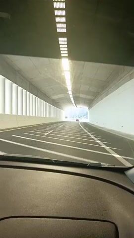 The video shows the interior of a car as it drives through a tunnel and then exits onto an open road. The tunnel is well-lit with overhead fluorescent lights, and the road is marked with white lines. Sunlight streams in from the tunnel's exit, illuminating the road ahead. Outside the tunnel, the car is on a highway with mountains in the background. A speed limit sign, showing a crossed-out motorcycle, is visible on the right side of the road. The car's dashboard display shows radio information. The video captures the transition from the enclosed space of the tunnel to the expansive outdoor environment.