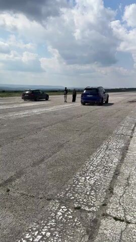 The video shows an open, paved runway with two cars racing, kicking up dust, and two people standing nearby, appearing to observe. The car on the left is darker and seems to be losing ground to the blue car on the right. The sky is partly cloudy, with some distant hills visible in the background. There are painted white lines on the runway, which appears to be old and somewhat cracked.