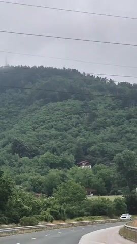 This is a landscape video shot from a vehicle moving on a road. The video shows a heavily wooded hillside under a cloudy sky. Two houses are visible nestled among the trees on the slope of the hill. A white pickup truck is driving on the road in the foreground. Power lines are visible against the sky.