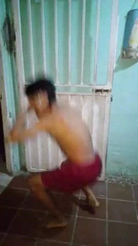 A shirtless person wearing red shorts crouches repeatedly in front of a white door with vertical bars. The person has dark hair and is facing away from the camera at an angle, with their movements blurry. The floor is tiled in reddish-brown squares, and the wall is a light teal color. A small, light-colored object is attached to the upper right corner of the wall.