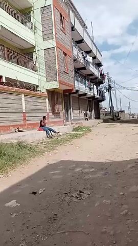 A person sits on a low concrete wall next to a street. The street is unpaved with dirt and debris. Tall buildings line the street. A small red car is parked next to a fence. A person stands near the car next to blue barrels. Laundry hangs from balconies on the buildings. Power lines run overhead.