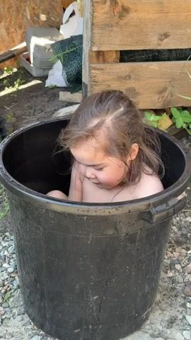 A young child with dark hair is seated inside a large black plastic bucket outdoors. Initially, the child looks down into the bucket. An adult voice is heard. The child then looks up, smiles, and picks up a blue and orange toy water gun, holding it in front of them. The ground around the bucket is dirt and gravel, with a wooden structure and some greenery visible in the background.