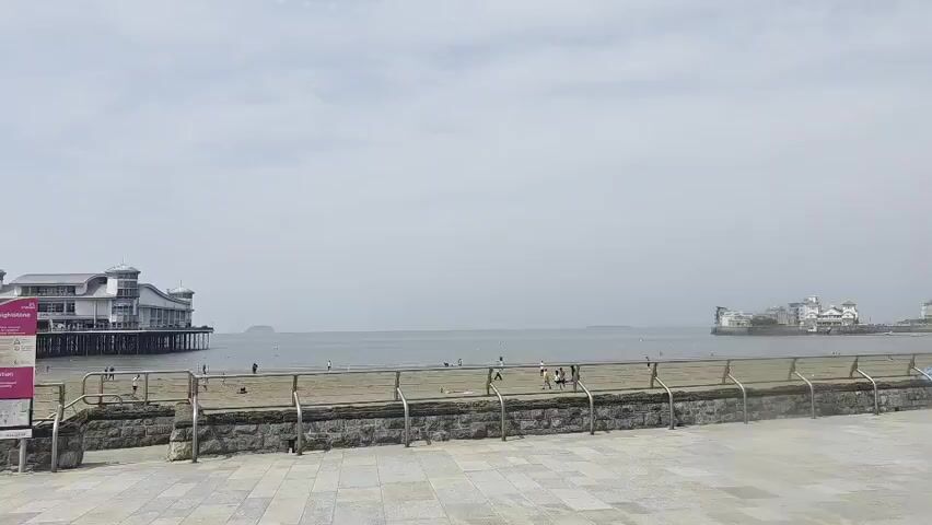 The video shows a view of a beach and the sea. In the distance, there are two piers. One pier, on the left, is larger and more elaborate with a white building at the end. The other pier, on the right, is smaller and appears to be an amusement pier with a few structures on it. The beach itself is sandy and appears to have a few people walking or standing on it. There is a metal fence and a stone wall separating the beach from a paved area in the foreground. The sky is overcast, and the water is calm. A lamppost is visible in the foreground, partially obstructing the view.
