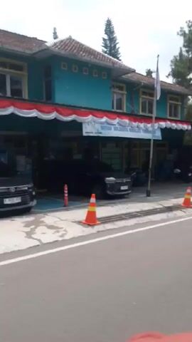 The video shows a street scene in Indonesia. The camera moves past a turquoise building decorated with red and white bunting and an Indonesian flag. There are vehicles parked in front of the building. Next, a yellow and blue pickup truck is seen parked in front of a blue and white PLN (Indonesian electrical company) sign. The camera continues to move, and a man in black clothing is seen walking alongside a white fence decorated with red and white fabric. Trees are visible in the background. The footage seems to be taken from a moving vehicle.