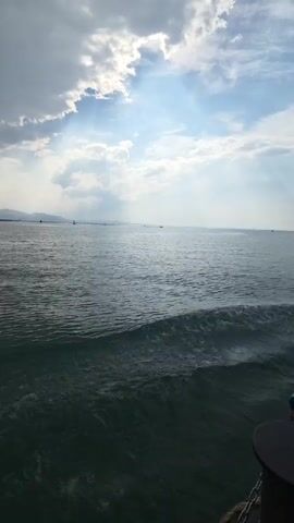 A video captured from a boat shows a wide expanse of water under a partly cloudy sky. Sunlight breaks through the clouds, casting rays onto the sea. Small waves ripple across the water's surface. In the distance, several boats are visible, including a sailboat and a speed boat. The coastline is faintly visible on the horizon. The perspective suggests the camera is in motion, possibly as the boat travels through the water.