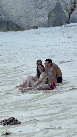 Three people are sitting on a sandy beach. Two women are sitting side-by-side, facing the camera. A man is sitting behind them, with his arms around the two women. All three people are covered in sand. The woman on the left is smiling. The woman in the middle has her eyes closed and is leaning her head against the man's head. The man has short dark hair and tattoos on his arms. The woman on the left is wearing a blue bikini top, and the woman in the middle is wearing a yellow bikini top. The man is wearing dark swimming trunks with a floral print. In the background, there is a large rock and some vegetation. The sand is white and has streaks of darker sand. There is a dark, lumpy object in the foreground. The overall impression is one of relaxation and camaraderie.