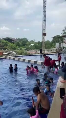 In a wide, bright, daytime shot, a group of children are in a swimming pool with blue water, some wearing swimsuits of various colors, while an adult male in a red shirt and light-colored shorts stands among them. The pool has a set of square stepping stones going across.  Behind the pool is a lush, green landscape with trees and some buildings. A tall metal structure, possibly a tower, is on the right side of the frame. At the edge of the pool, another adult can be seen partially, holding a phone, seemingly recording the scene. The children are moving around, interacting with each other and the adult in the red shirt. The sky above is partly cloudy.