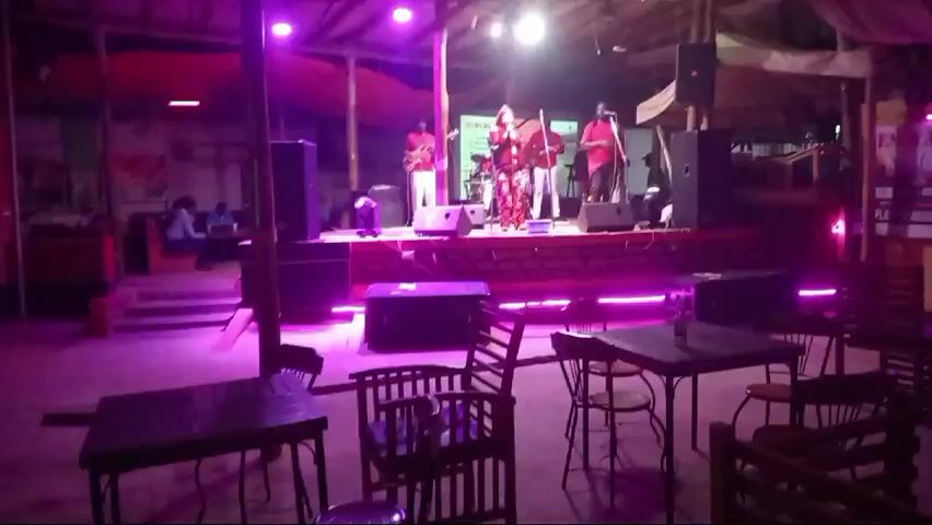 The video depicts a live band performing on a stage in a dimly lit, open-air bar or restaurant setting. The stage is adorned with purple LED lights along its base, and spotlights illuminate the performers. The band consists of five members: a female vocalist, two guitarists, a drummer, and a bassist. The vocalist stands center stage, dressed in a patterned outfit. The other band members are generally attired in red shirts and white pants. Two individuals are seated at a table in the left background. The foreground features several empty dark tables and chairs, suggesting the establishment is not very crowded. The building's structure is partially exposed, with visible wooden beams and a tarp-like ceiling. There is a vertical sign with yellow and white letters on the right side of the frame. The lighting alternates between shades of purple, blue, green, and white, creating a colorful ambiance. Throughout the video, a person wearing a black jacket and white pants is seen walking in the vicinity of the tables in the foreground.