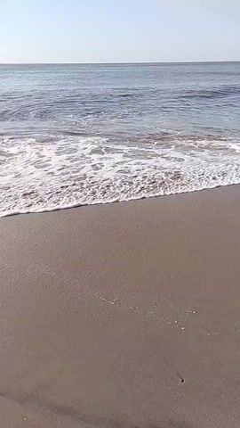 This video shows a serene beach scene. The camera is positioned low, close to the sand, capturing the gentle lapping of waves onto the shore. The water is a calm blue, with white foam trailing as the waves recede. The sand is a muted brown, appearing damp near the water's edge. In the distance, the horizon is visible where the sea meets a clear sky. There are no people or significant objects visible in the frame, focusing solely on the natural elements of the beach.