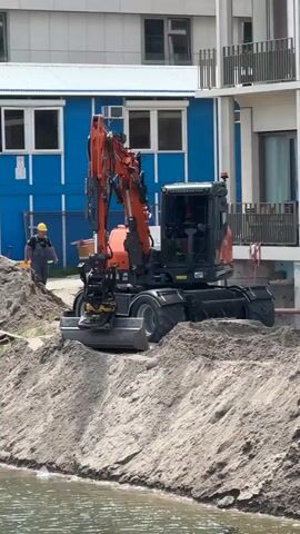 The video begins with a close-up of an orange excavator on a construction site, with a person in a yellow hard hat and blue overalls walking in the background. The excavator is positioned near a pile of dirt and a body of water. The scene then pans out to reveal a wider view of the construction site, showing more buildings under construction, including cranes and apartment buildings. There are also some houseboats docked along the canal. The weather appears to be overcast. The video focuses on the construction activity and the urban environment.