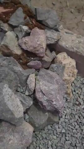 The video shows a close-up, shaky view of rocks and dirt. The rocks are of various sizes and colors, including gray, brown, and pink. Some of the rocks are covered in moss or dirt. There are also some reddish-brown pieces of brick scattered among the rocks. The ground appears to be dry and dusty. The camera moves slowly, panning across the scene. In the background, a dark fence or railing can be seen. The lighting is dim, suggesting it may be dusk or a shaded area. There are no people or animals visible in the video.