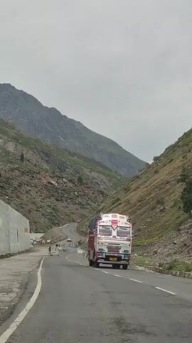 The video shows a truck on a winding road through a mountain pass. The truck is red and white and is driving on the right side of the road. The road is paved and has white lines to mark the lanes. The mountains on either side of the road are steep and rocky, with some vegetation. The sky is overcast. There are a few other cars on the road in the distance.