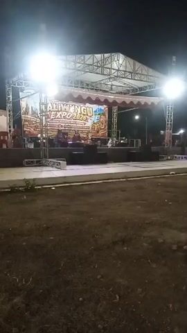 A nighttime outdoor stage is set up with a banner that reads "TALIWUNGU EXPO 2023." There are bright stage lights illuminating the performance area. A band or group of musicians is visible on stage, seated behind instruments. Speakers are set up in front of the stage, and various pieces of stage equipment are present. The area in front of the stage appears to be a dirt or gravel area. The overall scene suggests an outdoor event or concert.