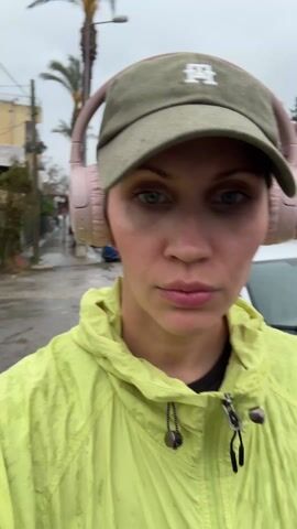 A woman in a yellow hooded jacket and a green baseball cap with a white logo is walking down a city street. She has pink headphones on her head. Cars and palm trees are visible in the background. The sky is overcast, and the street appears wet. The woman is looking at the camera.