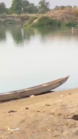 The video shows a calm river scene with a narrow wooden boat beached on a sandy bank in the foreground. In the background, a person is paddling a similar boat across the river. The opposite bank has some vegetation. The sky is overcast.