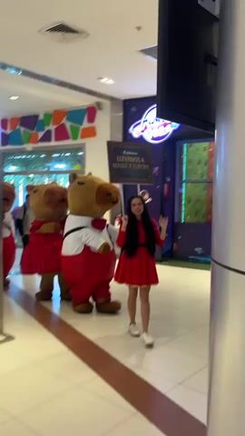 Inside what appears to be a brightly lit shopping mall, three capybara-like mascots, all dressed in red-and-white attire, stand beside a young woman in a matching red dress and white sneakers. She is holding a sign that reads "Llevamos a Mama a Europa." The girl is waving at the camera and smiling.  To the left is a display for "Ajedrez Gigante". Behind them is the entrance to “Mundo”.