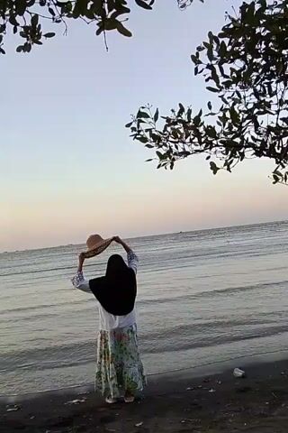 A person stands on a dark sand beach facing the ocean and adjusts a wide-brimmed straw hat. The person is wearing a white top with patterned sleeves and a long, floral-patterned skirt, along with a black hijab. The ocean has gentle waves, and the sky is a pale, light blue with a hint of pink near the horizon. Tree branches with green leaves hang down from the top of the frame, partially obscuring the sky. There is a piece of white trash on the beach to the right of the person.