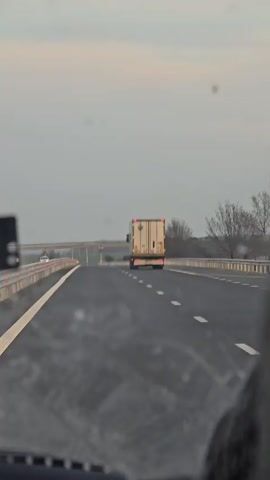 The video, captured from inside a moving vehicle, shows a view of a highway with a large trailer truck in the distance, traveling in the same direction. The highway is paved with asphalt, marked by dashed white lines. A bridge is visible in the background, spanning over a grassy field. The sky is overcast. A hint of the dashboard of the recording vehicle can be seen in the lower portion of the frame.