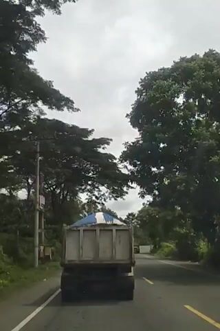 The video shows a dump truck on a road, viewed from behind. The truck is loaded with what appears to be sand or gravel, covered with a blue and white tarp. The truck is in the right lane of a two-lane road, with trees lining both sides. The sky is overcast. The truck has a white sign on its side and a yellow object is visible on the truck's rear. A second, smaller yellow vehicle is visible further down the road in the distance. Motorcycles are also visible on the road. The truck appears to be moving.