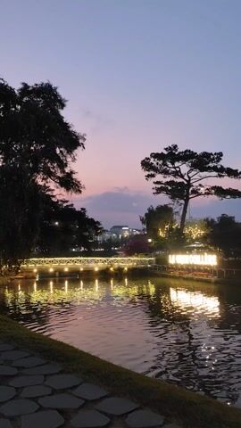The video shows a serene lake at dusk with a bridge adorned with lights crossing over it. The water reflects the lights and the illuminated sign of “LA CRÉMAILLÈRE” on the lake's edge. Silhouetted trees frame the scene against a dusky purple sky. A paved stone path borders the lake. The environment suggests a peaceful, possibly tourist-oriented, setting.
