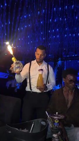 A server in a white shirt and suspenders holds a bottle of orange liquid and a lit sparkler over a table. A man wearing sunglasses and a patterned shirt is seated at the table. In the background, the ceiling is adorned with numerous small blue lights. The room appears to be a nightclub or bar.