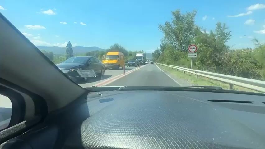 The video shows a view from inside a car dashboard looking out at a highway with heavy traffic. A yellow van is visible on the left side of the road. Several cars and trucks are in the lanes ahead, creating a traffic jam. There are trees and foliage on the sides of the road and a blue sky with some clouds. A road sign on the right indicates a 1000m distance. The car is stopped or moving very slowly in traffic.