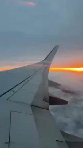 The video shows the view from an airplane window, focusing on the wing of the plane against a backdrop of clouds and a vibrant sunset. The sky transitions from a blue-gray hue to a fiery orange where the sun dips below the horizon. The clouds appear dense and fluffy. The airplane wing is primarily white, with visible panel lines and control surfaces.
