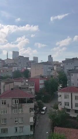 The video shows an aerial view of a city. In the foreground, there are several apartment buildings, some with red tiled roofs and others with gray facades. Trees are visible in front of and between the buildings. In the background, taller modern buildings, including skyscrapers and a minaret, are visible against a partly cloudy sky. The sun is shining, casting light on the scene. A white van is parked on a street between the buildings in the midground. The video pans slightly to the right.