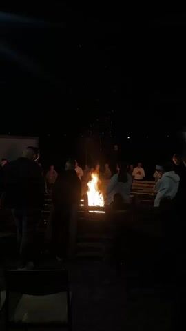 A group of people are gathered around a bonfire at night. The fire is burning brightly and sparks are rising into the dark sky. Most people are standing and facing the fire. Some are holding their hands up. The scene is dimly lit by the fire.