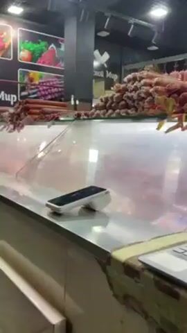 The video shows a laptop sitting in front of a display case filled with sausages. The screen of the laptop shows what appears to be a sales or inventory program. There is a payment terminal to the left of the laptop. Various types of sausages are hanging above the display case. The lighting is bright and artificial. The background shows other items for sale, possibly fruits or vegetables.