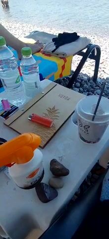 The video opens with a close-up view of a table on a pebble beach. On the table are two plastic water bottles, a pack of cigarettes, a red lighter, a wooden menu with a pineapple imprint and the word "HOYAH" on it, a cup with a straw and a beverage, and a bottle of sunscreen. In the background, there are several people in the ocean, and a few boats are visible in the distance. The video pans slightly to the left, revealing a person's legs and feet resting on a striped towel on a chair. The sky is blue with scattered white clouds. The waves are gentle, and the water is clear. The overall scene depicts a relaxed day at the beach.