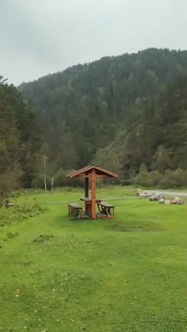 The video opens with a view of a wooden picnic shelter with a table and benches situated in a grassy clearing. In the background, a densely wooded hill rises, with a road visible in the mid-ground and a cluster of rocks to the right. The scene appears overcast. The camera then pans and moves, showing a dense green foliage. Finally, the camera focuses on two individuals, a man and a woman, sitting on a wooden structure by a body of dark water. The man, wearing a blue cap and a backpack, is seated to the left, while the woman, in a white jacket, is to the right. Several water bottles are placed on the wooden surface between them. The surrounding area is overgrown with green vegetation.