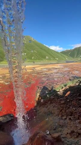 A waterfall cascades into a vibrant, reddish-orange pool of water in a mountainous landscape. The water in the pool is a striking red, with some areas appearing more yellowish or brown. The surrounding terrain is rocky and earthy, with green vegetation on the slopes of the mountains in the background. The sky is a clear, bright blue, suggesting a sunny day. The scene evokes a sense of natural wonder and perhaps a touch of the surreal due to the unusual coloration of the water. The time of day appears to be midday, given the bright sunlight. The location is identified as Ketrisi, Georgia.