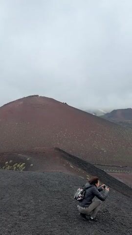 A woman with a backpack squats on a dark, rocky slope, holding a camera, pointed at a vast volcanic landscape under a cloudy sky. The landscape includes a large, reddish-brown hill, a winding road, and patches of vegetation, suggesting a volcanic environment with ongoing ecological development. The distant hills are partially obscured by low-hanging clouds. The road passes through a forested area. The sky is overcast.
