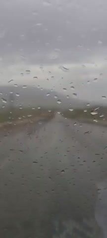 The video captures a first-person perspective from inside a moving vehicle, looking out through a rain-splattered windshield. Numerous raindrops adhere to the glass, blurring the distant view. Ahead, a wet, paved road stretches into the distance, appearing somewhat desolate with rolling hills or mountains visible on the horizon, shrouded in a misty, overcast sky.

As the vehicle travels, a white car briefly comes into view on the right side of the road, moving in the same direction as the camera's vehicle before quickly receding. The overall weather condition is rainy, with a grey, cloudy sky indicating an overcast day. The scene suggests a journey through a rural or semi-rural landscape. The provided context places this scene in Kan Mehtarzai, Pakistan. There is no visible text in the video. Background audio consists of what appears to be instrumental music playing, possibly from a car radio.