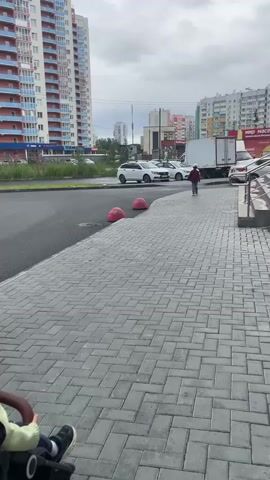 The video captures a street scene in Chelyabinsk, Russia. The foreground features a gray paved sidewalk with a child in a stroller on the left. The child is wearing a light green hooded jacket and dark pants with white-soled sneakers. In the midground, a person walks along the sidewalk, wearing a red cap, plaid shirt, and dark pants. They are walking away from the camera towards a large white truck. Several white cars are parked along the road.

In the background, tall, multi-story apartment buildings dominate the skyline. One building on the left is particularly prominent, with a red and white facade. Other buildings, varying in color and design, are visible further in the distance. A large white truck with a trailer is positioned on the road, and the rear of another white vehicle is visible on the right. Red, spherical bollards are placed along the edge of the sidewalk.

The sky is overcast, suggesting a cloudy or possibly rainy day. The lighting is even, indicating it is likely daytime, perhaps late morning or afternoon. The overall atmosphere appears to be that of a typical urban environment, with residential buildings, infrastructure, and pedestrian activity. There are no explicit signs of strong emotions or a specific event, suggesting a candid shot of everyday life.

Visible text in the image includes "МИР МАССА" (MIR MASSA) on a red banner on one of the background buildings. A sign with the number "30" is visible on the building to the right of the frame.