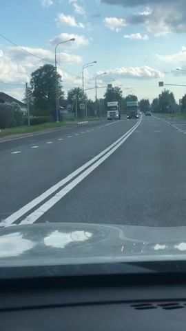 The video shows a first-person perspective from inside a moving car driving on a multi-lane asphalt road in Opolie, Russia. The road has a double solid white line separating opposing traffic and multiple lanes marked with dashed white lines further ahead. Overhead, streetlights and yellow pedestrian crossing signs are visible. On the left side of the road, there are trees and some buildings, suggesting a semi-rural or suburban setting.

Several vehicles are seen driving towards the camera. Initially, a white truck is in the left lane, and a green Scania truck, appearing to carry logs, is in the right lane. These trucks are passed by the recording vehicle. Later, a white coach bus passes on the right. Other distant cars are also visible on the road.

The sky is partly cloudy but bright, indicating daytime. Reflections of the car's dashboard and windshield are visible in the lower foreground of the frame. There is no clearly readable text on signs or vehicles within the video.
