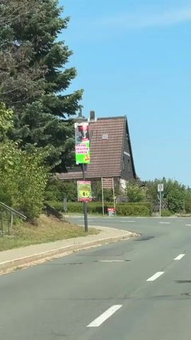 The video shows a rural road with a curve ahead. On the left side of the road, there is a tall evergreen tree, a sidewalk, and a lamppost with two signs attached. The top sign is red and white and appears to be an advertisement. The bottom sign is green and white with pictures of children. Further down the road, there is a house with a brown tiled roof and yellow siding. There are bushes and trees surrounding the house. A speed limit sign is visible on the right side of the road. The sky is blue and clear. The video is shot from a moving vehicle.