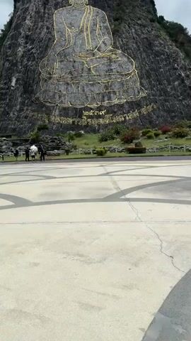 A medium shot captures a colossal Buddha image etched in gold onto a sheer rock face, dominating the frame against a cloudy sky. The Buddha is depicted in a seated position with intricate details in its robes and headdress. Below the image, Thai script is inscribed on the rock face. In the middle ground, a paved plaza with geometric patterns leads towards the rock. A group of approximately five people, appearing to be tourists, are visible in the lower left quadrant of the image, standing on a grassy area near the base of the rock. They are casually dressed, with some holding umbrellas. Lush green vegetation and some flowering bushes dot the landscape around the plaza. The overall scene conveys a sense of awe and reverence for the monumental artwork.
