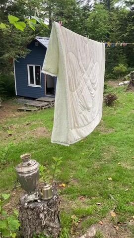 A metal samovar and teapot sit on a tree stump in the foreground. A large, light-colored quilt hangs from a clothesline in the middle ground, with a small blue building to its left. The background is filled with lush green trees and foliage, creating a natural, outdoor setting. The quilt is white with a subtle patterned texture. The building has a dark roof and blue walls with a white-trimmed window. The overall scene suggests a peaceful, rustic environment, possibly a backyard or garden.