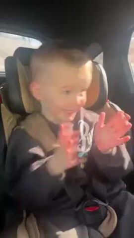 A young boy wearing a car seat is shown clapping his hands. The boy is wearing a dark gray sweatshirt, and a beige vest-like strap is covering his chest. He is looking to his right as he claps his hands together quickly. Sunlight can be seen coming in through the car window, and the boy’s fair skin appears to be lightly flushed with color. The car seat is dark with tan accents, and the background is slightly blurry.