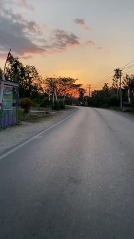 A low-angle, first-person perspective video shows a silver minivan driving on a paved road during sunset. The road curves to the left, with a dirt shoulder on the left side and trees lining the right side. Power lines are visible overhead. The sky is filled with orange and pink clouds, and the sun is setting behind a cluster of trees in the distance. The minivan is the only vehicle visible in the frame and it drives away from the camera.