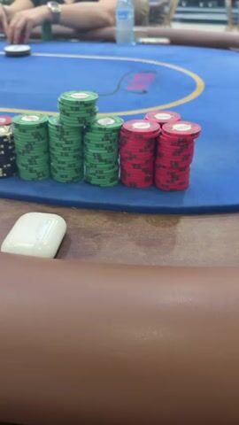 The video captures an ongoing poker game from a first-person perspective at the table. Several individuals are seated around a large blue felt poker table, which is adorned with gold lines and rectangular cutouts. Stacks of poker chips, primarily in green and red colors, are visible on the table, indicating different players' chip counts. Some stacks are tall, suggesting significant bets or accumulated winnings.

Playing cards, mostly white with red suits (likely diamonds or hearts), are laid out on the blue felt, possibly showing community cards or a player's hand. In the foreground, a white AirPods case rests on the brown wooden edge of the table. Hands of players can be seen occasionally, moving chips or interacting with cards. The atmosphere appears casual, with a water bottle and other small items on the table in the background. The lighting suggests an indoor setting, possibly a casino or a dedicated game room. No specific text is clearly legible on the chips or cards.