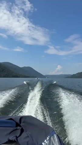 A view from the stern of a moving boat looking out at a calm, wide lake. In the foreground, the white wake of the boat is prominent. Further out on the lake, several other boats are visible, also moving and creating their own wakes. The lake is surrounded by densely forested, mountainous terrain. The sky is blue with scattered white clouds. The overall impression is a pleasant day spent on the water.