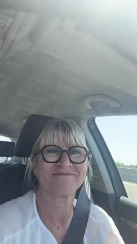 A woman with blonde hair and glasses is sitting in the driver's seat of a car. She is wearing a white shirt and a seatbelt. The car appears to be on a highway, as indicated by the road and trees visible outside the window. The sun is shining brightly, causing some glare in the video. The woman is looking at the camera and smiling, and her facial expression changes slightly throughout the short clip.