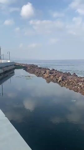 The video shows a body of water, likely the ocean, with a rocky breakwater in the foreground. The water is calm with small waves. On the right side of the breakwater, a wooden pier extends into the water, with a small shelter at the end. The sky is cloudy and the overall scene is a coastal landscape.